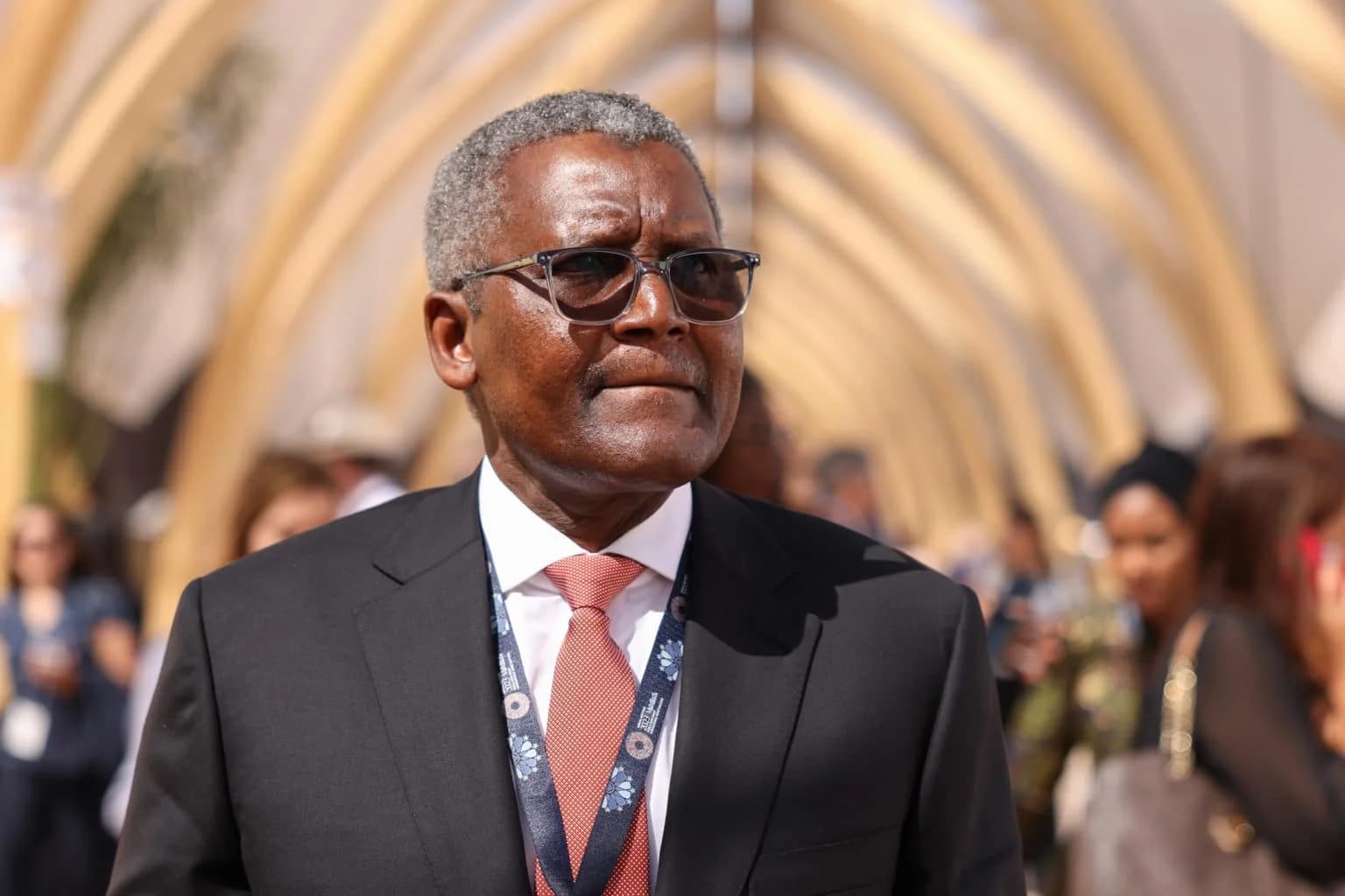Aliko Dangote wearing sunglasses and a conference lanyard, walking through an archway