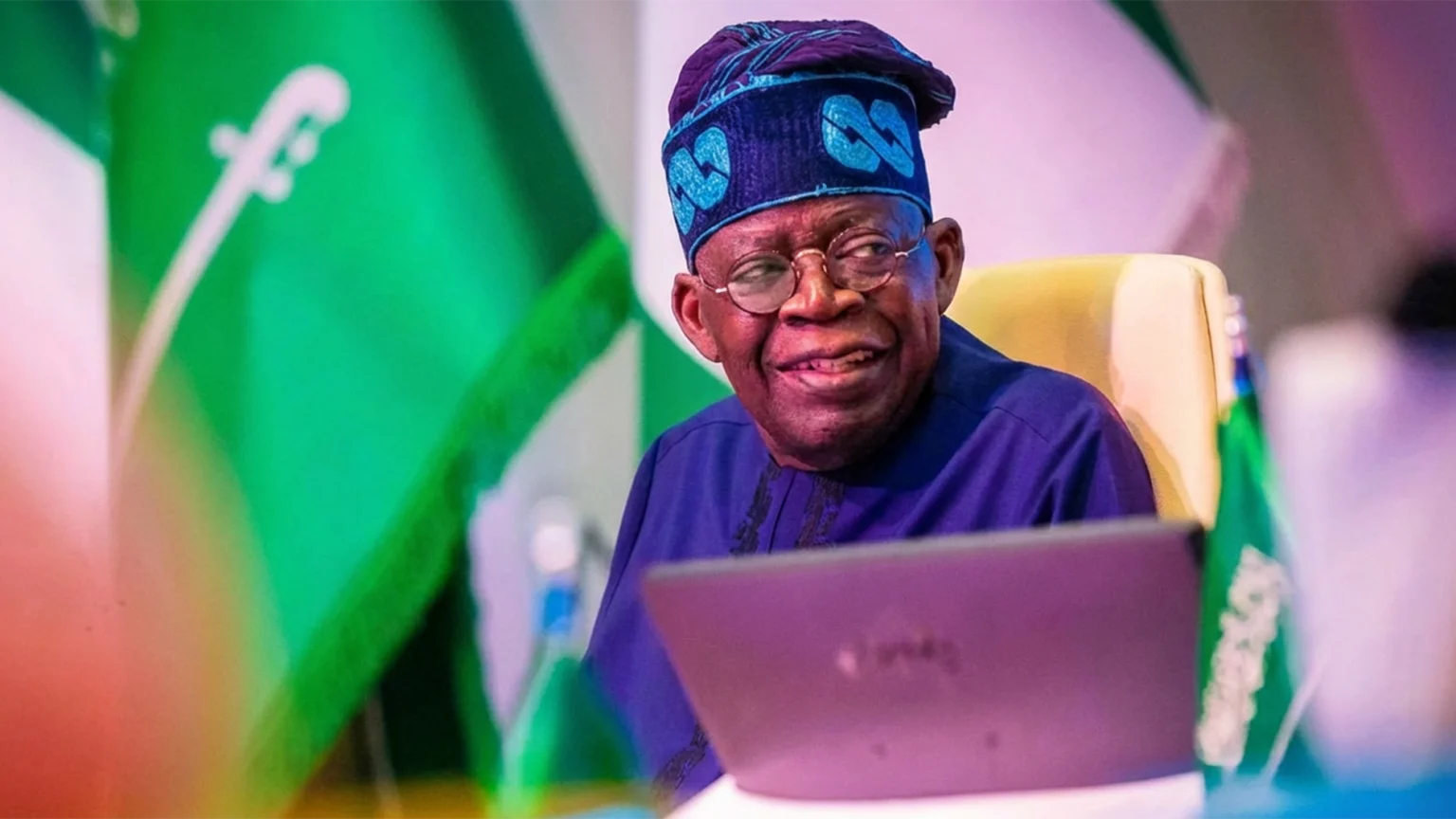 Nigerian President Bola Tinubu looking at a laptop screen, flanked by national flags