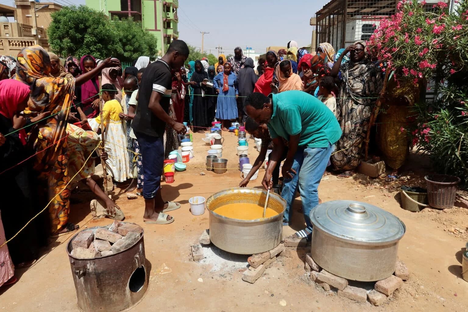 A crowd lines up with buckets and containers around large cooking pots of food in a Sudanese street